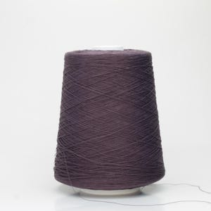 May include: A large cone of dark purple yarn, tightly wound, sits on a white base. The yarn is a solid color, with a slight sheen, and appears to be made of a soft material. A strand of yarn extends from the cone.