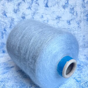 Puede incluir: Una madeja de hilo de color azul claro con una textura peluda. El hilo est&aacute; enrollado alrededor de un tubo de cart&oacute;n con una etiqueta de cinta azul.