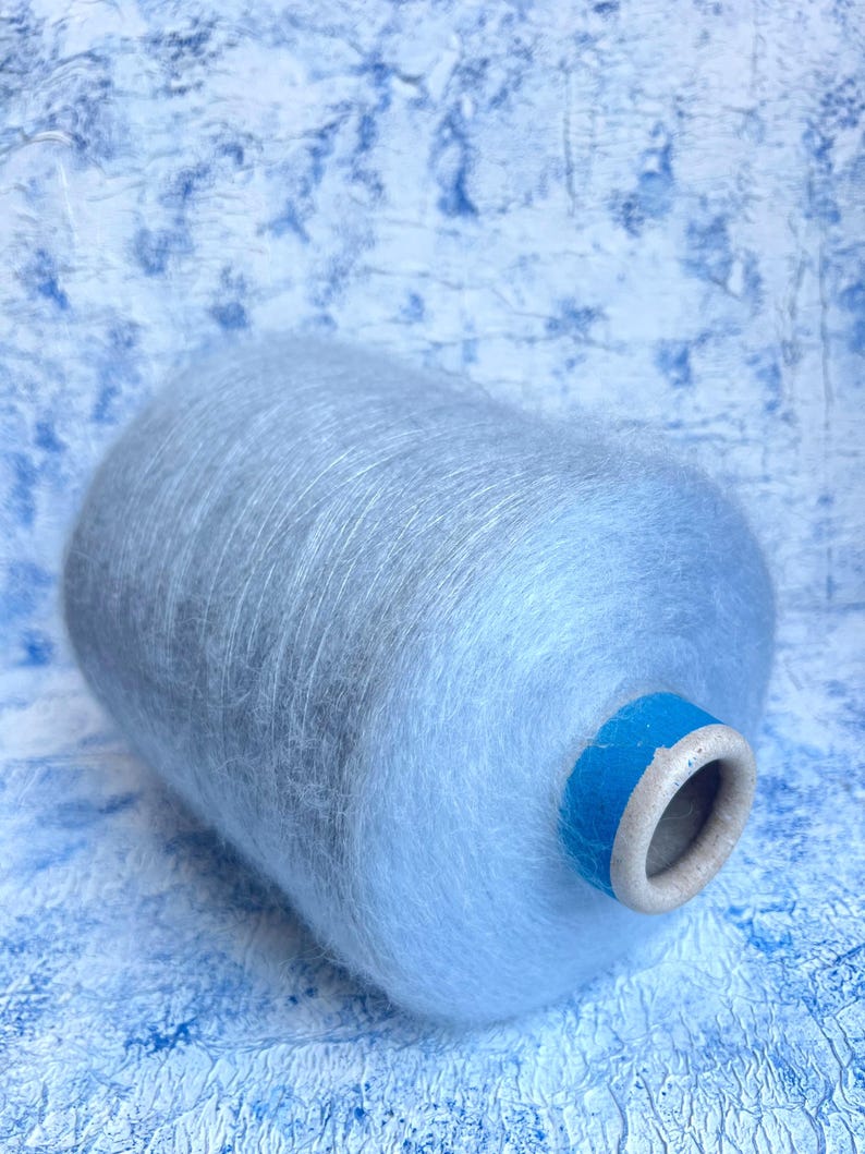 Puede incluir: Una madeja de hilo de color azul claro con una textura peluda. El hilo est&aacute; enrollado alrededor de un tubo de cart&oacute;n con una etiqueta de cinta azul.