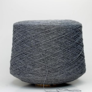 May include: A large cone of grey yarn, tightly wound. The yarn is a medium weight, suitable for knitting or crochet projects. The background is plain white, highlighting the yarn's texture and colour. The yarn is likely to be sold by the gram.
