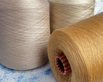 May include: Three spools of yarn in varying shades of beige and tan. The yarn is tightly wound on cardboard cores. The colors range from light beige to a warm, golden tan. The background is a textured, light blue and white surface.