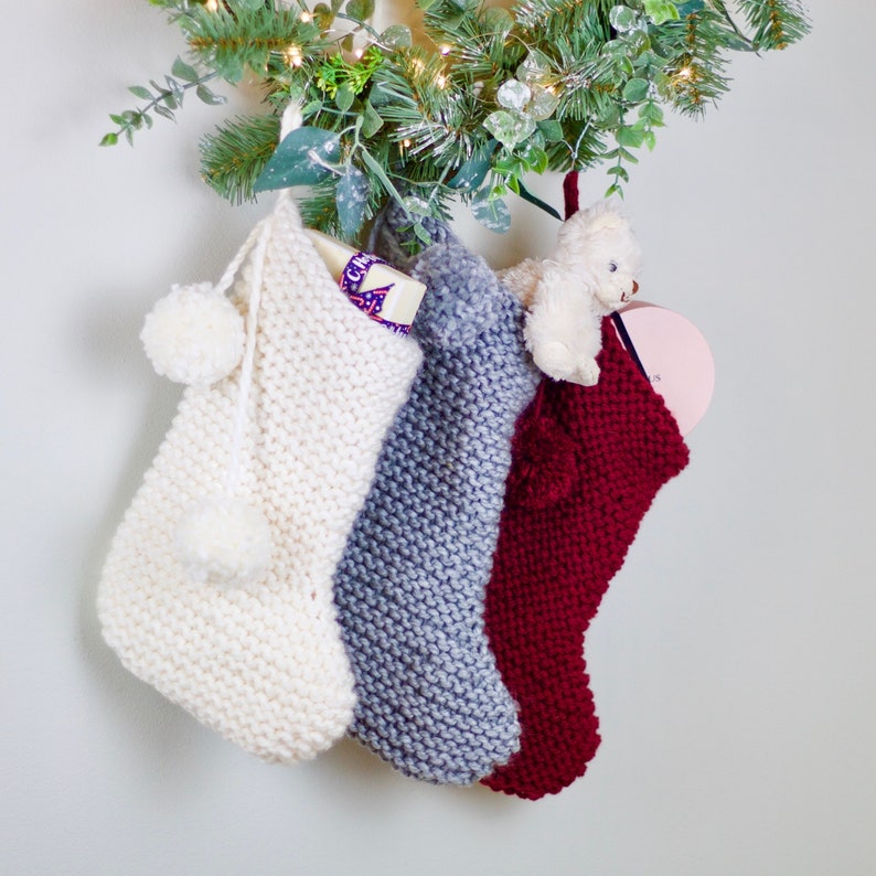 May include: Three knitted Christmas stockings hanging from a green garland. The stockings are white, gray, and red. The white stocking has a white pom-pom on the top and a small white gift inside. The gray stocking has a white pom-pom on the top. The red stocking has a white pom-pom on the top and a small stuffed animal inside.