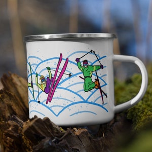 May include: White enamel mug with a blue and white wave pattern. The mug features a colorful illustration of three skiers on a snowy mountain.