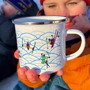 May include: White enamel mug with a silver rim and a curved handle. The mug features a blue wave pattern and colorful skiers in action. The skiers are wearing various colors of ski suits and are holding ski poles. The mug is being held by a person.