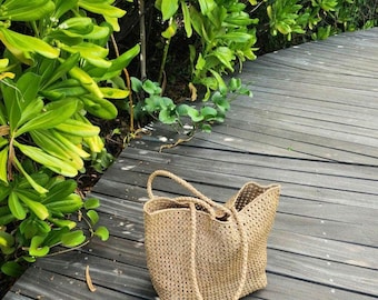 Könnte beinhalten: Eine große, gewebte Tragetasche aus Naturfasern. Die Tasche hat eine hellbraune Farbe und eine lockere, offene Webart. Die Tasche liegt auf einem Holzsteg.