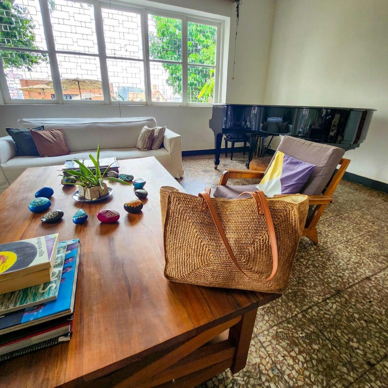 May include: A woven straw tote bag with brown leather handles sits on a wooden coffee table. The table also holds a stack of books, painted stones, and a small plant. A white sofa and a piano are in the background.
