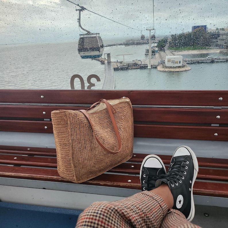 May include: A woven brown tote bag with leather handles sits on a wooden bench. Black high-top sneakers with white soles and a star logo are visible. The background shows a cable car and a coastal view on a rainy day.