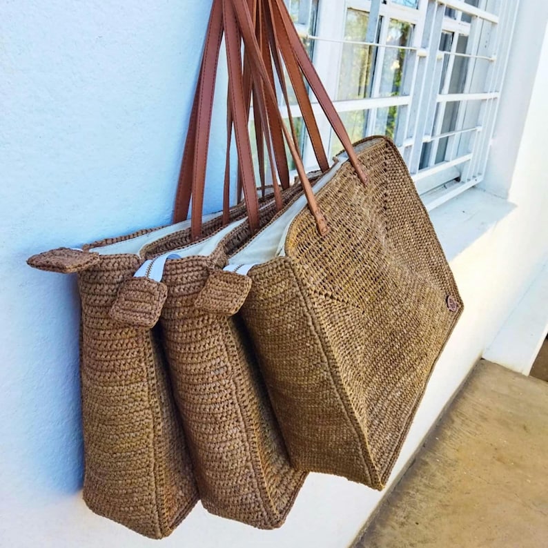 May include: Several woven tote bags with brown leather handles hang against a white wall. The bags are a natural tan color, with a rectangular shape and a woven texture. The handles are a rich brown, and the bags appear to be lined with white fabric.
