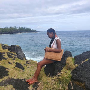 May include: A woman sitting on a rock near the ocean, holding a large, woven straw tote bag. She wears a white top, denim shorts, and orange sandals. The bag is rectangular with a woven pattern and brown leather straps.