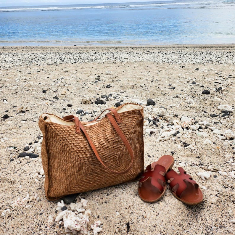 May include: A woven brown tote bag with leather handles and a pair of brown leather sandals rest on a sandy beach. The ocean is visible in the background, creating a relaxed, summery scene.