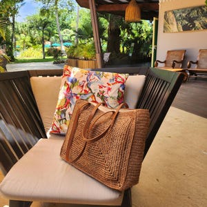 May include: A woven brown tote bag with leather handles sits on a chair with a floral pillow. The bag has a rectangular shape and a textured surface. The pillow features a colorful floral pattern with red, blue, and white flowers.