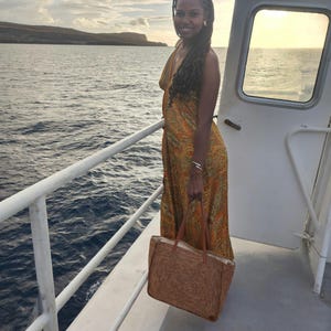 May include: A woman wearing a long, orange paisley print dress stands on a boat, holding a woven tote bag with brown leather handles. The background shows the ocean and a distant island under a cloudy sky.