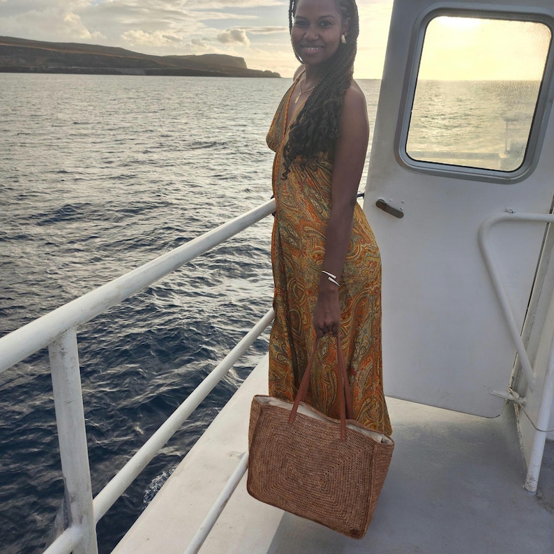 May include: A woman wearing a long, orange paisley print dress stands on a boat, holding a woven tote bag with brown leather handles. The background shows the ocean and a distant island under a cloudy sky.