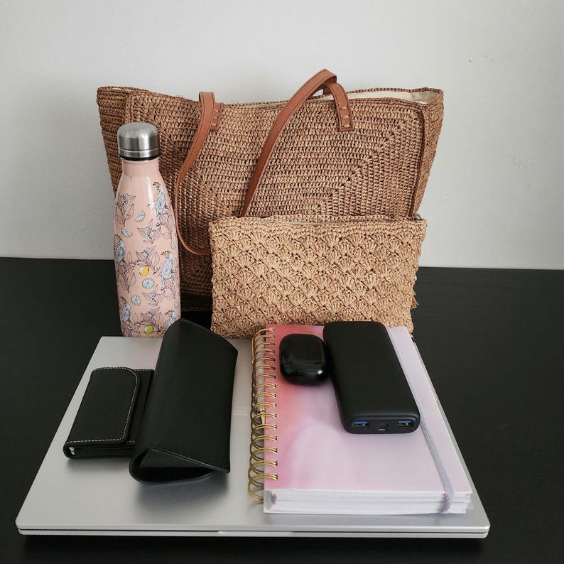 May include: A collection of everyday carry items, including a woven tote bag with leather handles, a matching clutch, a pink floral water bottle, a laptop, a notebook, a power bank, a wallet, and wireless earbuds. The items are arranged on a black surface.