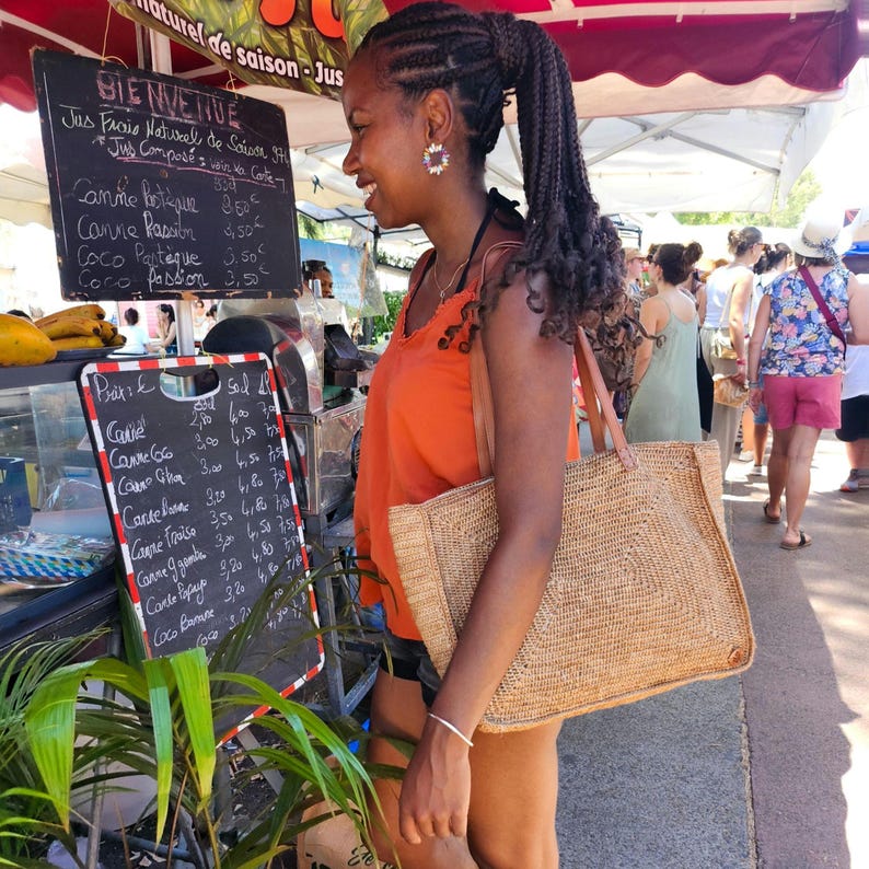 May include: A woman stands at a juice stand, holding a woven tote bag. The stand features a chalkboard menu with handwritten text in French. The woman wears an orange top and black shorts. The background shows other people and market stalls.