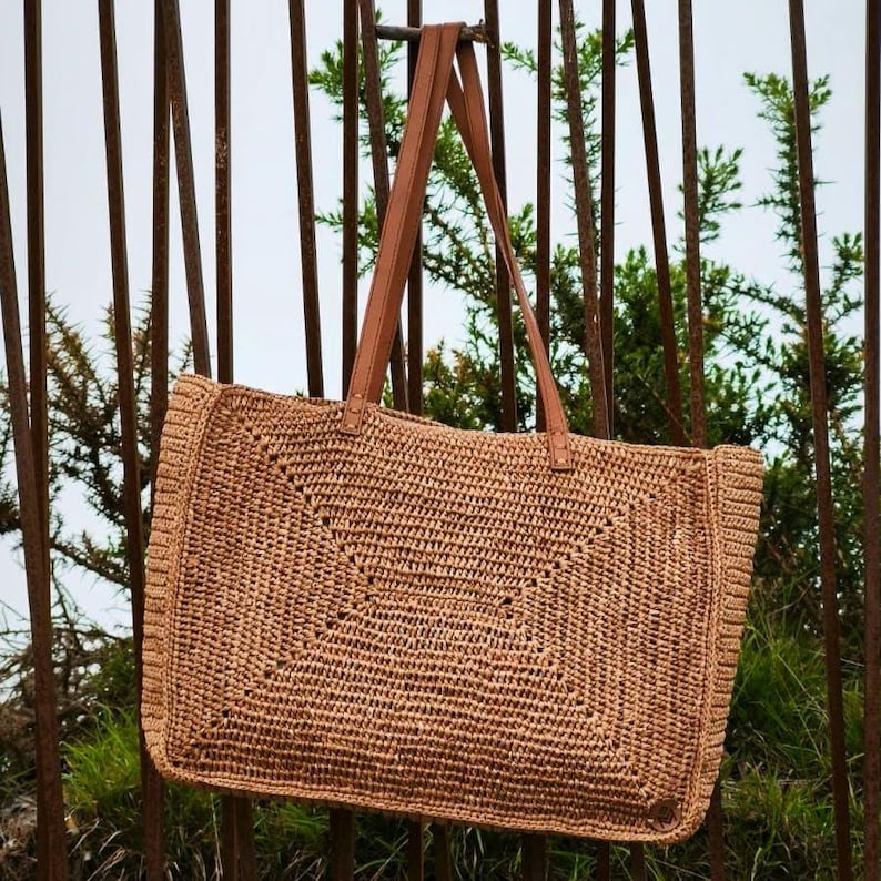 May include: A woven tote bag in a natural tan color, featuring a geometric design on the front. The bag has brown leather handles and is hanging against a background of metal bars and greenery. The bag is rectangular in shape.