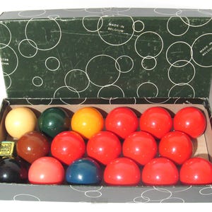 May include: A vintage box set of billiard balls. The set includes 15 red balls, along with balls in yellow, green, brown, black, and blue. The box is dark green with a white circle pattern and the text "MADE IN BELGIUM".