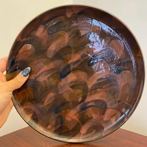 May include: A large, round ceramic plate with an abstract design in shades of brown, pink and blue. The plate has a glossy finish.