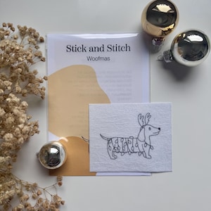 Op de afbeelding: Een knutselpakket met de tekst "Stick and Stitch Woofmas" met een teckel met geweien en kerstverlichting. De set bevat een bedrukt stoffen ontwerp, met gouden en zilveren kerstballen.