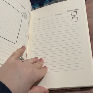 May include: Open notebook with lined pages and sections for campaign, character name, and session notes. The left page has a dotted grid and a compass graphic. The right page is open, showing the lined pages. The notebook is open, revealing the interior pages.