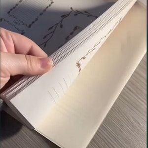 May include: A notebook with cream-colored pages is being opened, revealing the interior. The pages have a delicate floral design in a light brown color. The notebook is on a gray surface.