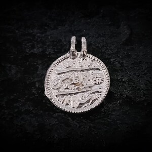 May include: A silver pendant with an intricate design and Arabic script. The round pendant has a detailed border and two loops at the top for a chain. The pendant is likely a piece of jewelry.