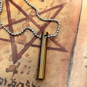 May include: A gold-colored, cylindrical pendant necklace with a ball chain. The pendant is a small, hollow tube. The background is a textured, tan surface with a faded pentagram design.