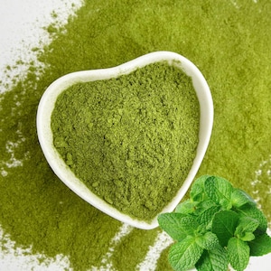 May include: A heart-shaped white bowl filled with bright green powder, surrounded by more of the same powder and fresh mint leaves. The powder has a fine texture, and the mint leaves are a vibrant green.