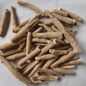 Puede incluir: Un mont&oacute;n de ra&iacute;ces de ashwagandha secas, de color marr&oacute;n claro. Las ra&iacute;ces son cil&iacute;ndricas, con una textura rugosa y longitudes variables. Est&aacute;n dispersas sobre una superficie clara, mostrando los tonos naturales y terrosos del producto herbal.