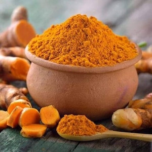 Puede incluir: Un cuenco de terracota lleno de polvo de cúrcuma de color naranja brillante. Raíces frescas de cúrcuma y trozos cortados están esparcidos alrededor del cuenco. Una cuchara de madera contiene un pequeño montón del polvo, mostrando el color y la textura de la especia.