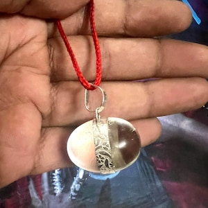 May include: A clear crystal pendant with a silver filigree design, hanging from a red cord.