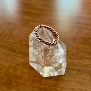 May include: A copper wire ring with a twisted design, resting on a clear quartz crystal.