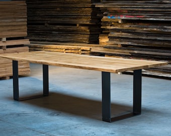 May include: A long, wooden dining table with a live edge top and black metal legs. The table is in a warehouse setting with stacks of wood planks in the background.