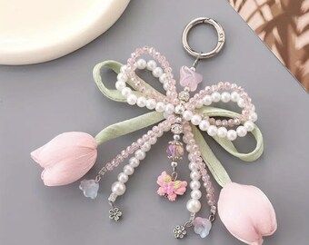 May include: A decorative keychain featuring a bow design made of white faux pearls and light pink beads. The keychain includes two light pink fabric tulip flowers, a pink butterfly charm, and a silver-tone clasp.