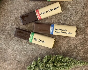 May include: Five chocolate bars with colorful wrappers and text in German. The wrappers have phrases like "dass Du meine beste Freundin bist!" and "für Dich!" The bars are arranged on a textured, neutral-toned surface.