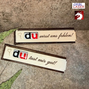 May include: Two wooden boxes with a brown border and a white interior. The boxes have a heart and the word "du" in red and black letters. The top box says "du wirst uns fehlen!" and the bottom box says "du tust mir gut!"