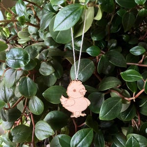 Op de afbeelding: Een houten kuiken ornament met een touwtje om op te hangen. Het kuiken is lichtbruin en heeft een simpel cartoon ontwerp.