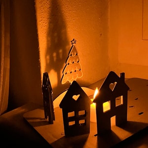 May include: A wooden Christmas village scene with a lit candle, a small house, and a tree. The houses are painted black and have white windows and hearts. The tree is painted brown and has a star on top.