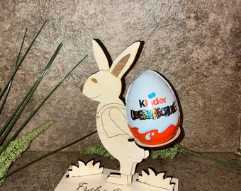 Könnte beinhalten: Holzfigur eines Osterhasen, der ein Kinder Überraschungsei hält. Der Hase steht auf einem Sockel mit dem Text "Frohe Ostern!"