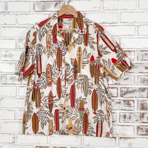 Puede incluir: Una camisa blanca de manga corta con botones y un estampado tropical de tablas de surf y hojas de palmera. La camisa tiene un esquema de color rojo, marrón y azul. La etiqueta de la camisa dice "Bishop St."