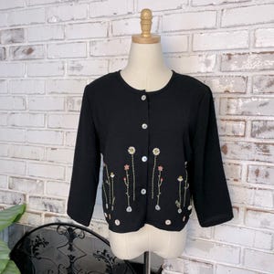 May include: Black cardigan with three-quarter sleeves and a round neckline. The button-down sweater features embroidered floral details in white, pink, and green. The cardigan is displayed on a mannequin against a white brick wall.