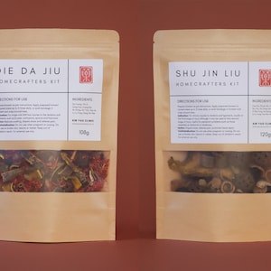 Könnte beinhalten: Zwei Kraftpapierbeutel mit transparenten Fenstern, beschriftet mit "DIE DA JIU" und "SHU JIN LIU HOME CRAFTERS KIT." Jeder Beutel enthält Kräuterzutaten. Der "DIE DA JIU" Beutel wiegt 108g, und der "SHU JIN LIU" Beutel wiegt 120g. Beide haben Anweisungen und eine Zutatenliste.