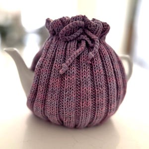May include: A purple and pink knitted teapot cosy with a drawstring closure. The cosy is made of a soft, textured yarn and is designed to keep tea warm.