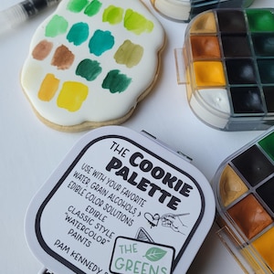 May include: A cookie decorated with watercolor paints in shades of green, yellow, and brown, alongside a cookie palette kit. The kit includes a palette with various colors and the text "THE COOKIE PALETTE".