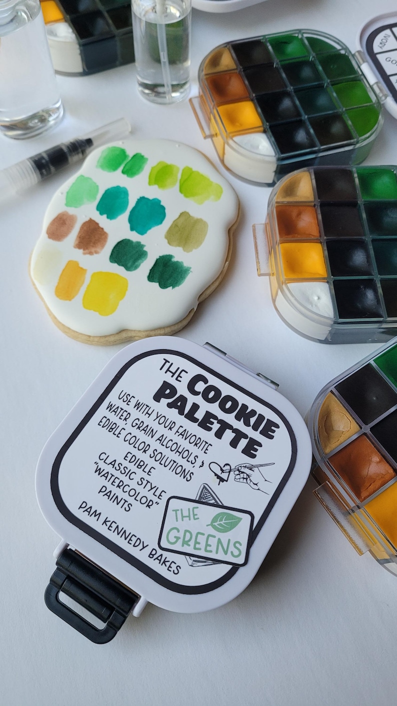 May include: A cookie decorated with watercolor paints in shades of green, yellow, and brown, alongside a cookie palette kit. The kit includes a palette with various colors and the text "THE COOKIE PALETTE".