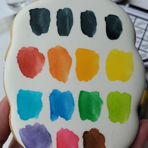 May include: A hand-held, cookie-shaped like a paint palette, decorated with white icing and colorful watercolor paint swatches. The swatches include shades of green, red, orange, yellow, blue, purple, pink, and brown.