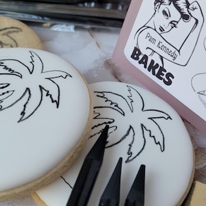 Peut inclure: Des biscuits ronds glac&eacute;s blancs avec des motifs de palmiers noirs et des crayons noirs. Un r&eacute;cipient rose avec une illustration de femme et le texte "Pam Kennedy BAKES" en arri&egrave;re-plan. Les biscuits ont un bord brun clair.