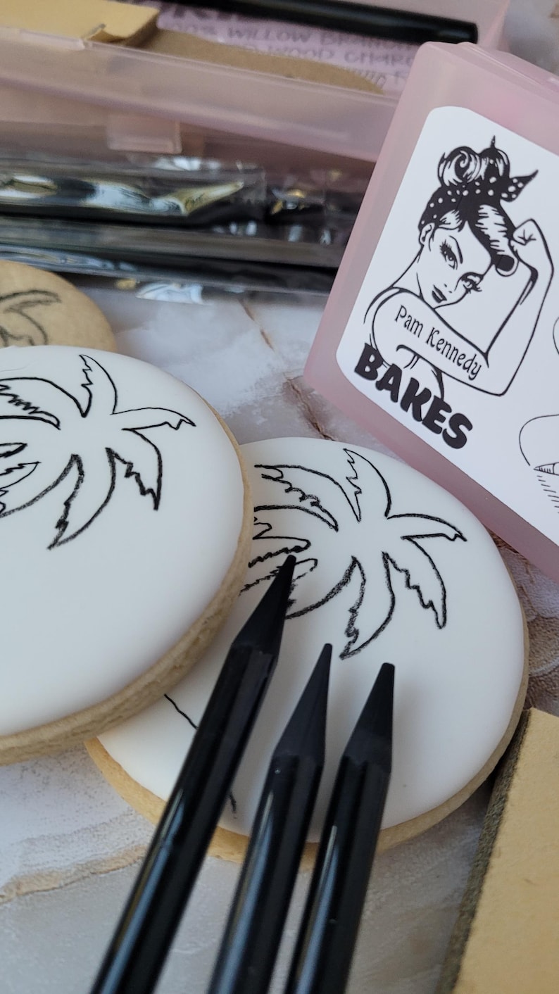 Peut inclure: Des biscuits ronds glac&eacute;s blancs avec des motifs de palmiers noirs et des crayons noirs. Un r&eacute;cipient rose avec une illustration de femme et le texte "Pam Kennedy BAKES" en arri&egrave;re-plan. Les biscuits ont un bord brun clair.