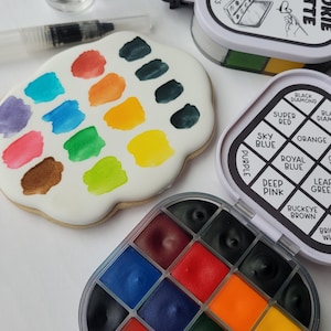 May include: A cookie palette with various edible watercolor paints. The palette includes a variety of colors, such as red, blue, green, and yellow. Also included are two bottles of liquid and a brush.