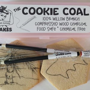 May include: A pink plastic box labeled "The Cookie Coal" with text and a drawing of a woman. Inside are black pencils and two cookies with drawings on them. The pencils are labeled "Cookie Coal".